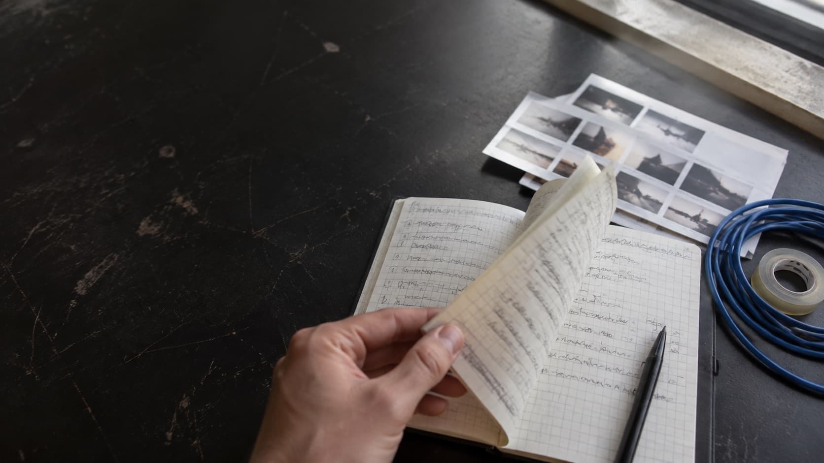 Mão a folhear um caderno de trabalho junto a provas fotográficas e um cabo azul